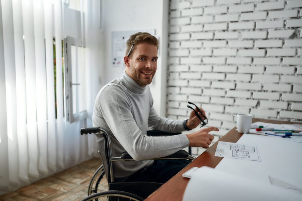 Hiring employees increases HR compliance responsibilities. This portrait shows a confident smiling young male seated in a wheelchair with a disability working with blueprints.